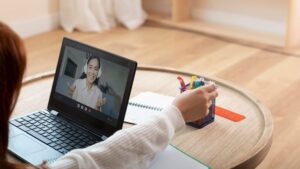 Na zdjęciu widoczna jest osoba siedząca przed laptopem na okrągłym, niskim stoliku. Głównym elementem jest otwarty laptop, na którego ekranie widać wideo połączenie. Na ekranie widać uśmiechniętą kobietę w słuchawkach.