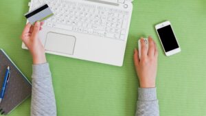Na zdjęciu widok z góry na biurko lub stół o jasnym, zielonym blacie. W centrum kompozycji znajduje się biały laptop z otwartą klawiaturą. Po lewej stronie widać dłoń trzymającą kartę płatniczą, co sugeruje dokonywanie transakcji online. Obok laptopa leży otwarty notes z niebieskim długopisem. Po prawej stronie, obok laptopa, jest biała myszka komputerowa. Z myszki korzysta druga dłoń. Obok myszki leży biały smartfon.