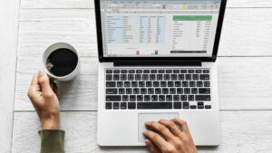
Na zdjęciu widać widok z góry na stół, na którym znajduje się otwarty laptop. Ekran komputera wyświetla arkusz kalkulacyjny, prawdopodobnie Microsoft Excel, z danymi i tabelami. Widać dwie dłonie osoby obsługującej komputer. Jedna dłoń, znajdująca się po lewej stronie, trzyma biały kubek z ciemnym napojem, najprawdopodobniej kawą. Druga dłoń spoczywa na panelu dotykowym (touchpadzie) laptopa. Stół jest wykonany z jasnego, bielonego drewna z wyraźnymi słojami.