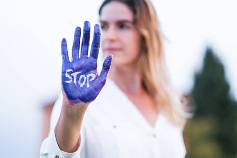 Na zdjęciu widzimy wyciągniętą dłoń pomalowaną na niebiesko z białym napisem „STOP” na środku. Kobieta w tle, ubrana w białą koszulę, jest lekko rozmyta, co kieruje uwagę na gest sprzeciwu.