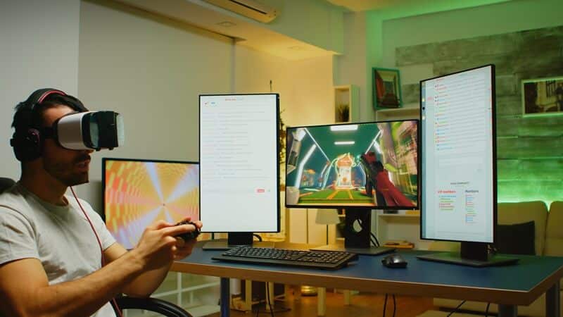 Na zdjęciu widzimy mężczyznę siedzącego przy biurku w nowoczesnym wnętrzu, ubranego w gogle VR i trzymającą kontroler do gier. Przed nim znajdują się trzy duże monitory: jeden pokazuje abstrakcyjny wzór, drugi scenę z futurystycznej gry, a trzeci interfejs czatu lub transmisji na żywo. Pomieszczenie jest stylowo oświetlone, z zielonymi akcentami świetlnymi i starannie dobranym wystrojem, w tym sofą, grafiką na ścianie i półkami.