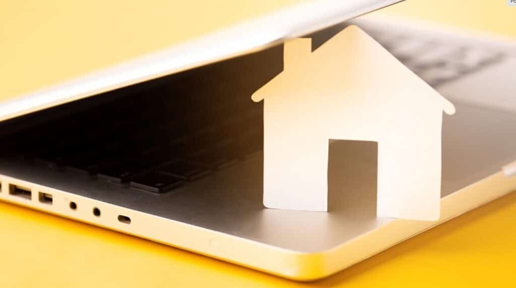 Na zdjęciu widzimy nowoczesną kompozycję symboliczną, przedstawiającą białą, papierową makietę domu częściowo wsuniętą pod ekran srebrnego laptopa. Ekran jest lekko przymknięty, co sprawia wrażenie, jakby dom był „wchłaniany” przez urządzenie. Tło w intensywnym, żółtym kolorze nadaje całości energii i kontrastu, podkreślając prostotę formy i czytelność przekazu.