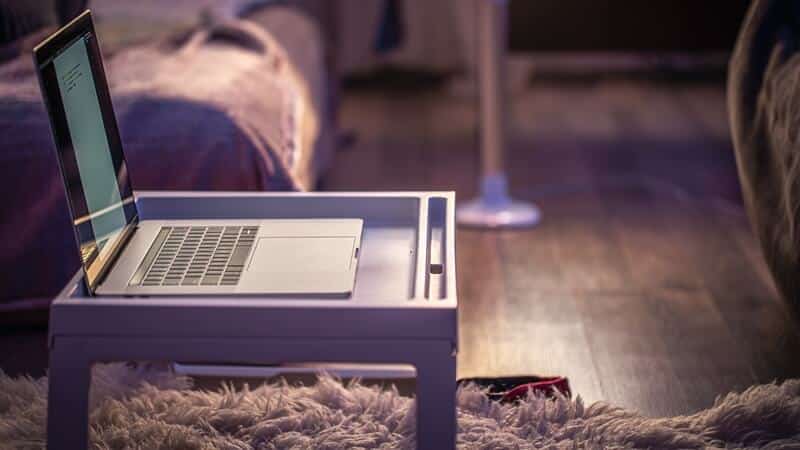 Na zdjęciu widoczna jest przytulna przestrzeń domowa, idealna do pracy lub nauki w spokojnej atmosferze. Na puszystym dywaniku stoi niewielki, biały stolik, na którym umieszczono otwarty laptop. W tle znajduje się łóżko z narzutą i poduszkami, a całość oświetlona jest miękkim, ciepłym światłem, prawdopodobnie z lampy stojącej widocznej w rogu pomieszczenia. Drewniana podłoga i stonowana kolorystyka wnętrza podkreślają domowy charakter miejsca, sprzyjający skupieniu, relaksowi i wygodnej pracy zdalnej.