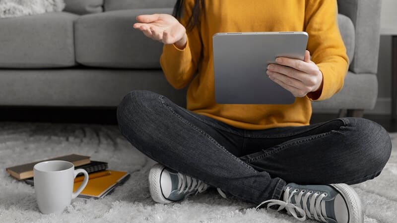 Na zdjęciu widzimy osobę siedzącą po turecku na dywanie, tuż przed szarą sofą. Ubrana jest w musztardowy sweter, ciemne jeansy i jasnoniebieskie sneakersy. W jednej ręce trzyma tablet, a drugą gestykuluje, co sugeruje, że uczestniczy w rozmowie online lub wideokonferencji. Obok niej leży biały kubek oraz kilka zeszytów — żółty i czarny — co wskazuje na domowe warunki pracy lub nauki. Całość tworzy swobodną, przytulną atmosferę sprzyjającą skupieniu i komunikacji zdalnej.