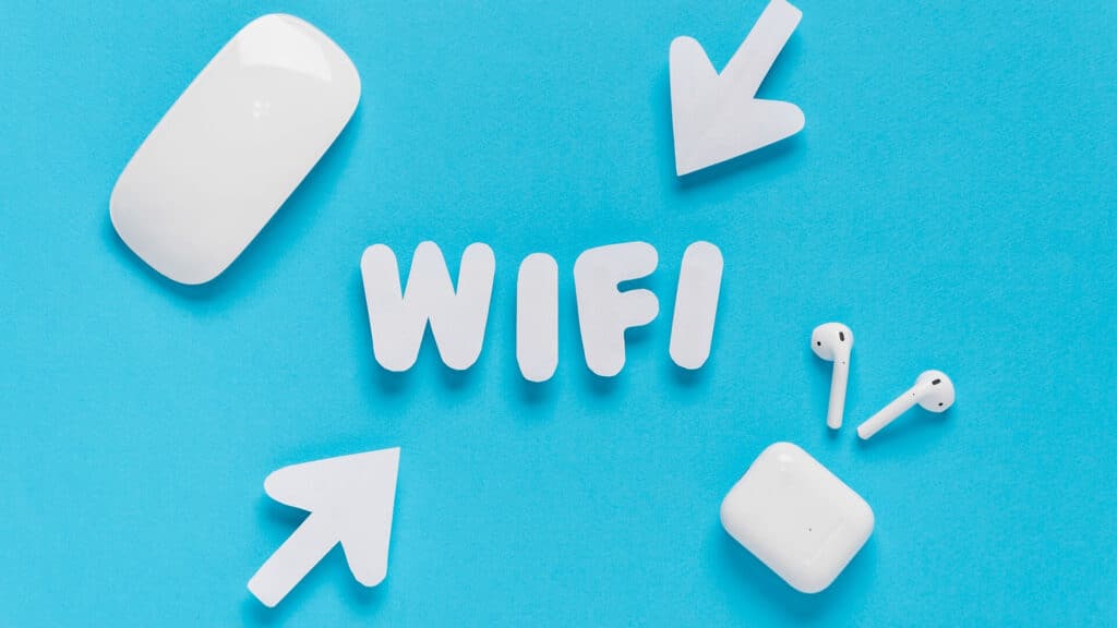 Na intensywnie niebieskim tle, które przywodzi na myśl technologię i nowoczesność, centralnie umieszczony jest biały, trójwymiarowy napis „WIFI”. Z trzech stron kierują na niego białe strzałki, podkreślając jego znaczenie jako centrum komunikacji. W prawym dolnym rogu znajdują się białe bezprzewodowe słuchawki z etui — eleganckie, minimalistyczne, jednoznacznie kojarzące się z mobilnością i wygodą.