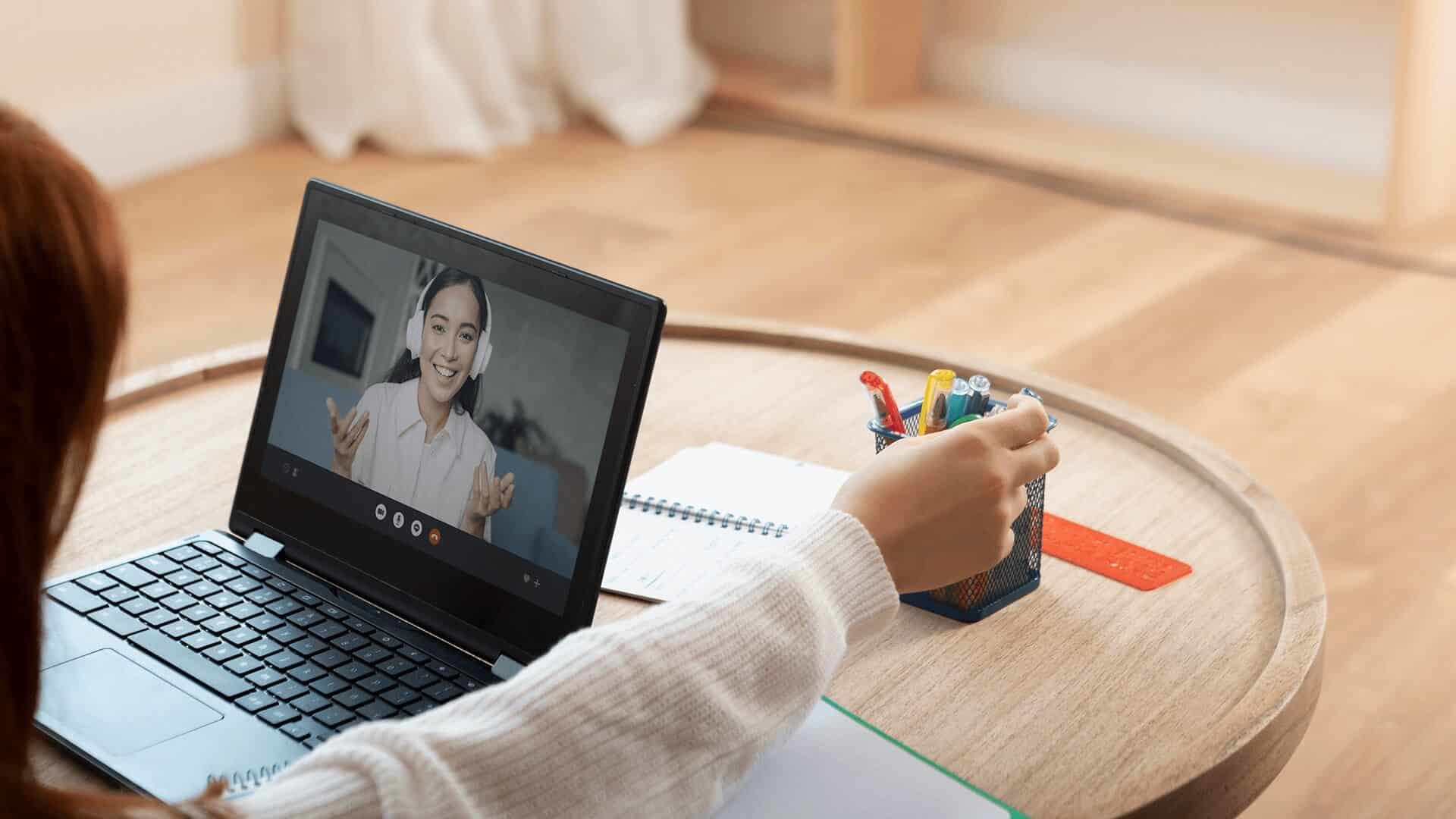 Na zdjęciu widzimy scenę pracy lub nauki zdalnej w domowym otoczeniu. Osoba, której rękę widać na pierwszym planie, siedzi przy okrągłym, drewnianym stole. Ma przed sobą laptopa, na którym widać ekran z trwającym połączeniem wideo – druga osoba na ekranie uśmiecha się i gestykuluje, prawdopodobnie prowadząc rozmowę lub lekcję online.

Na stole znajdują się także przybory do pisania: koszyk z kolorowymi długopisami i zakreślaczami oraz otwarty notatnik. Obok koszyka leży czerwona linijka. W tle widać jasną podłogę i zasłony, co nadaje przestrzeni przytulny, domowy charakter.