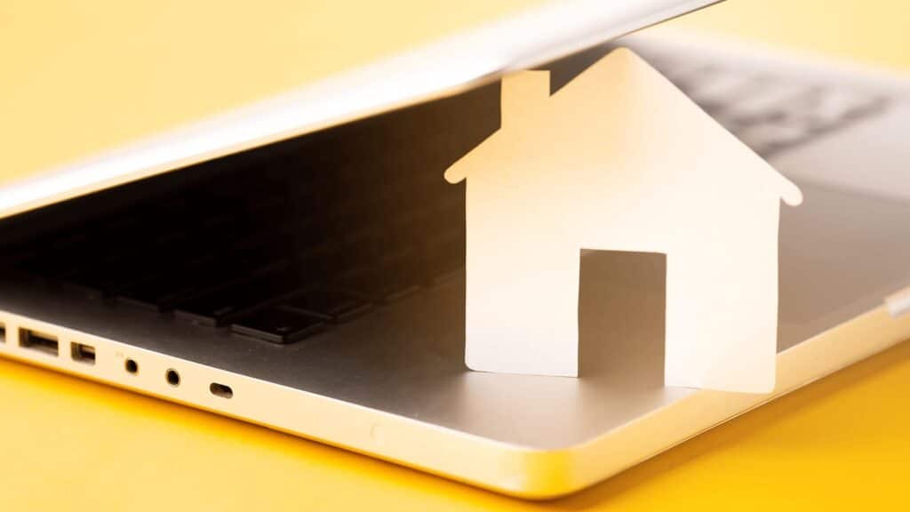 Na intensywnie żółtym tle, które przyciąga wzrok i nadaje całości nowoczesny, optymistyczny ton, znajduje się srebrny laptop z czarną klawiaturą. Jego ekran jest lekko przymknięty, a spod niego wystaje biały, papierowy wycinankowy domek — prosty, stylizowany, z wyraźnym dachem, kominem i drzwiami. Ten kontrast między fizycznym, dziecięco symbolicznym domkiem a cyfrowym urządzeniem tworzy sugestywną metaforę: dom przeniesiony do świata online.