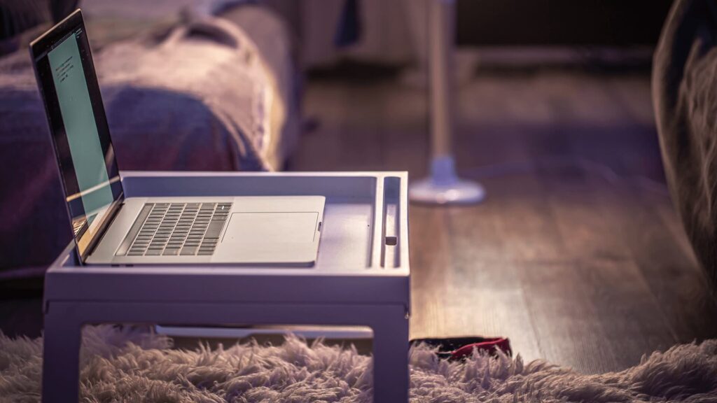 Zdjęcie przedstawia pokój, w którym na puszystym dywanie stoi niewielki, biały stolik lub taca, na której spoczywa otwarty laptop. W tle widać łóżko z miękkimi poduszkami i narzutą, a obok — smukłą lampę podłogową z jasnym kloszem. Światło w pomieszczeniu jest miękkie, przytłumione, jakby wieczorne, co nadaje całości przytulny, domowy klimat.