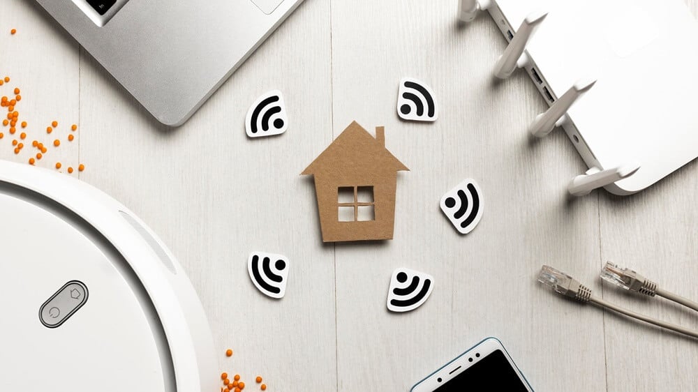 Na zdjęciu widać drewnianą makietę domu umieszczoną na jasnym, drewnianym blacie, otoczoną symbolami sieci Wi-Fi. Wokół domku znajdują się kilka białych elementów z czarnymi ikonami Wi-Fi, które symbolizują bezprzewodową łączność i dostęp do internetu. W kadrze widoczne są również fragment laptopa, smartfona, urządzenia sieciowego z antenami oraz przewody Ethernet.