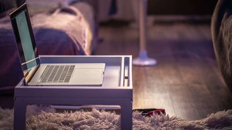 Na zdjęciu widać laptop ustawiony na niewielkim stoliku przy kanapie. Wokół znajduje się przytulne wnętrze z miękkim, włochatym dywanem i ciepłym, nastrojowym oświetleniem. Scena przedstawia komfortowe korzystanie z laptopa w domowym otoczeniu.